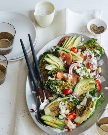 Grilled BLTA gem salad on a platter with serving utensils, plates, and drinks.