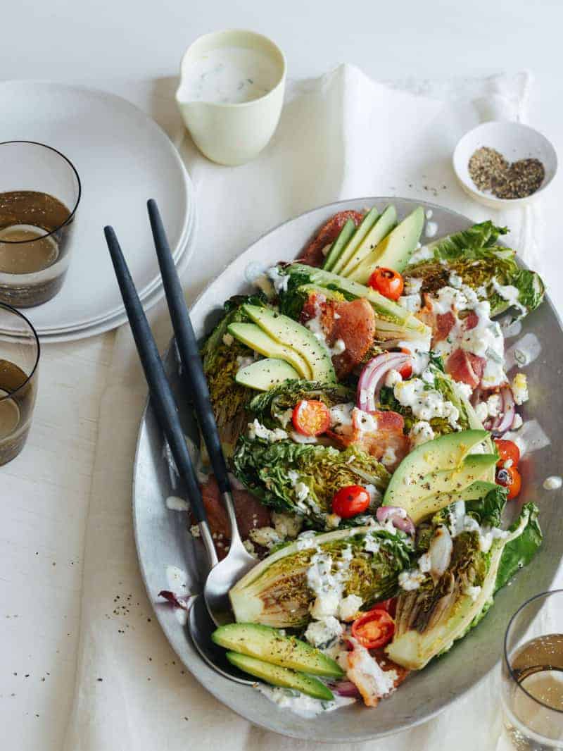 Grilled BLTA gem salad on a platter with serving utensils, plates, and drinks.