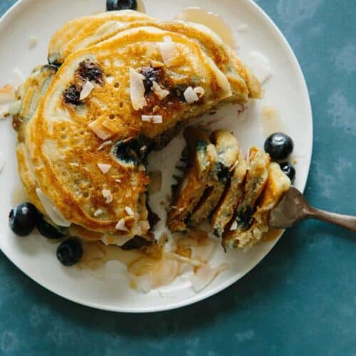 A plate of brown sugar, coconut, blueberry pancakes with a bite on a fork.