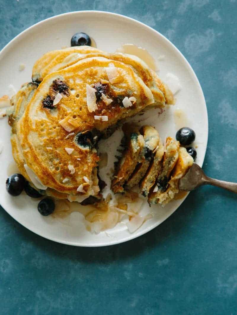 Brown sugar, coconut, blueberry pancakes drizzled in syrup with a bite on a fork.