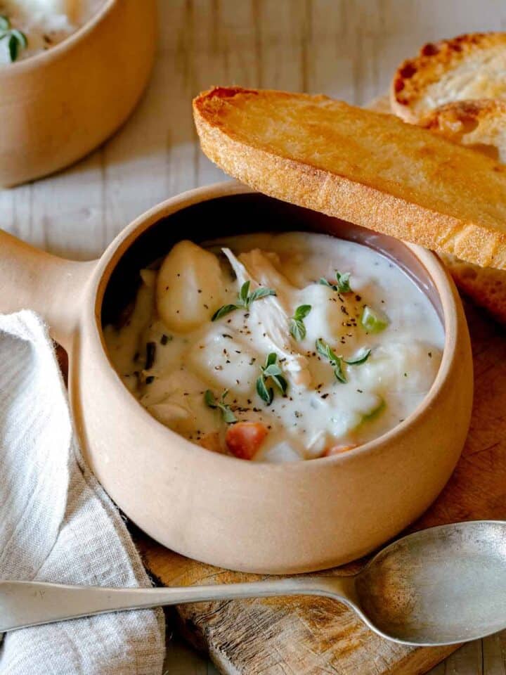 A soup bowl with handle of creamy chicken and gnocchi soup with bread and a spoon.