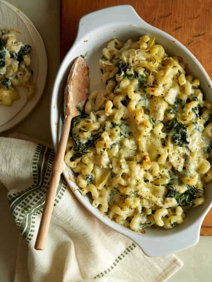 A baking pan of spinach artichoke mac and cheese with a wooden spoon.