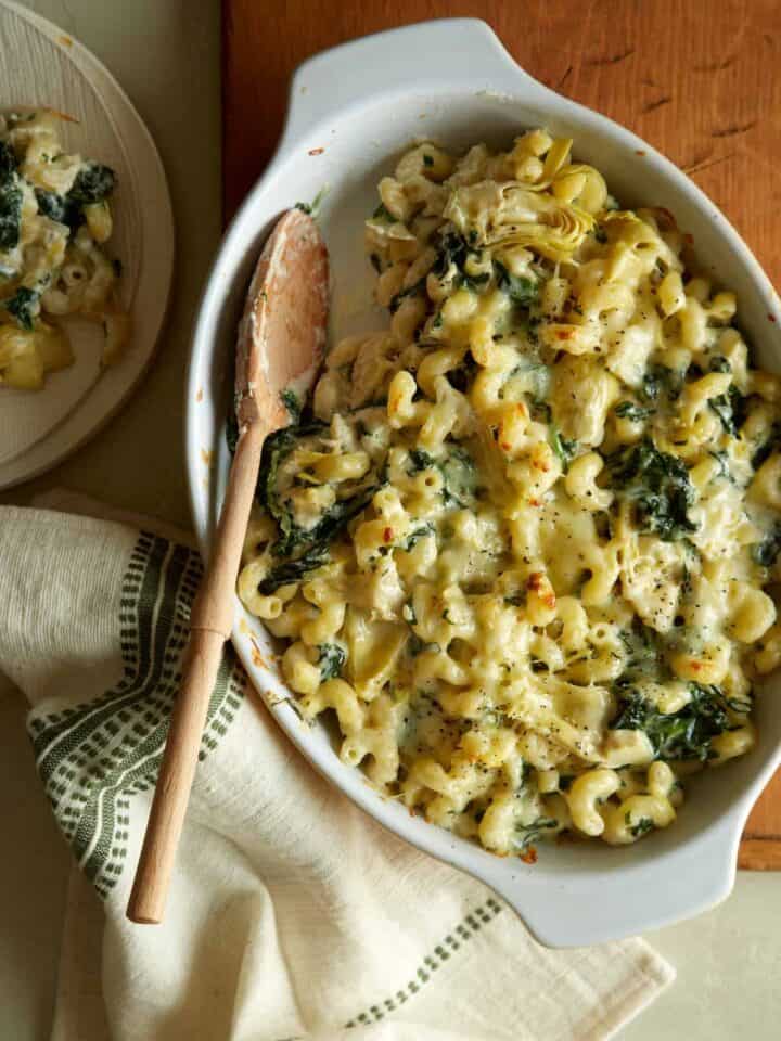 A baking pan of spinach artichoke mac and cheese with a wooden spoon.
