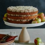 A whole apple cake with chai spiced buttercream on a cake stand with apples and plates.