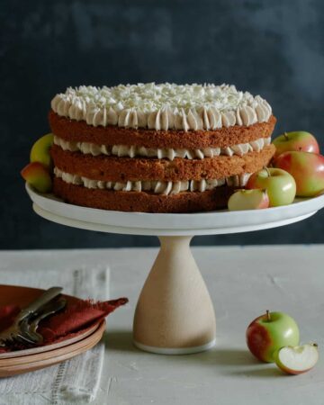 A whole apple cake with chai spiced buttercream on a cake stand with apples and plates.