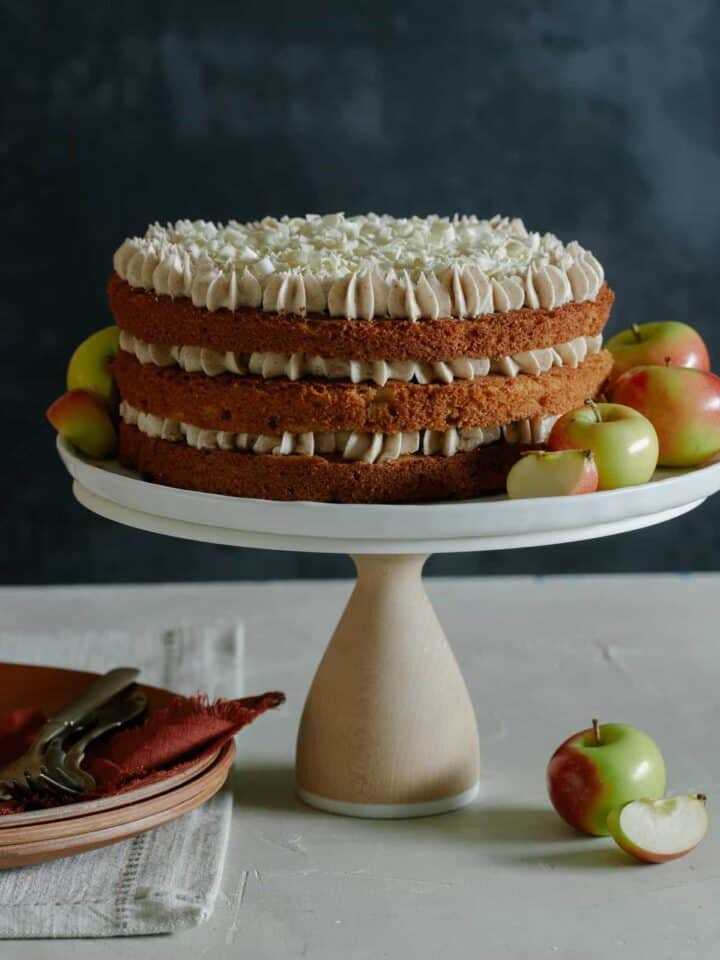 A whole apple cake with chai spiced buttercream on a cake stand with apples and plates.