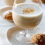 A close up of a glass of hot and creamy butterbeer on a plate with a cookie.