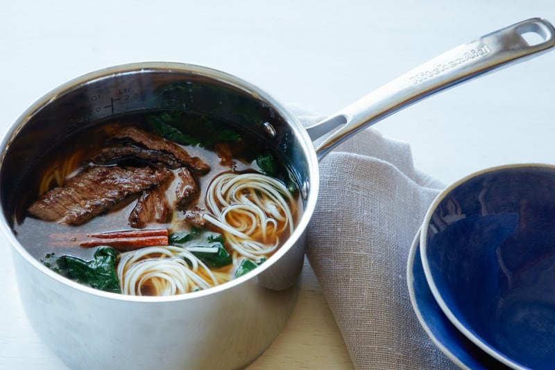 A pot of garlic ginger beef noodle soup with linens and blue bowls.