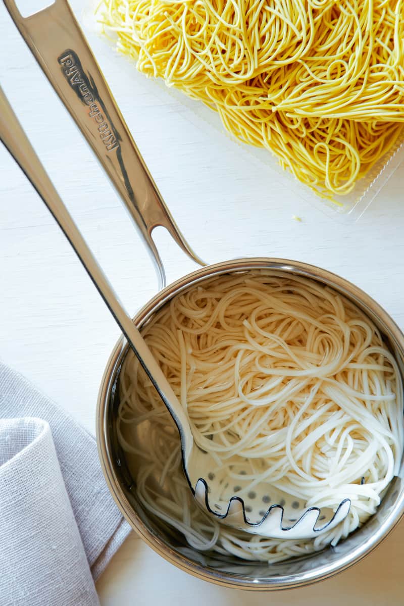 A pot of cooked noodles with a spaghetti fork and dry noodles to the side.