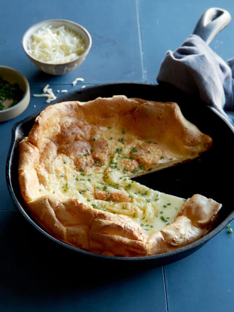 Herb and cheese dutch baby in a skillet with a slice taken out and a towel.