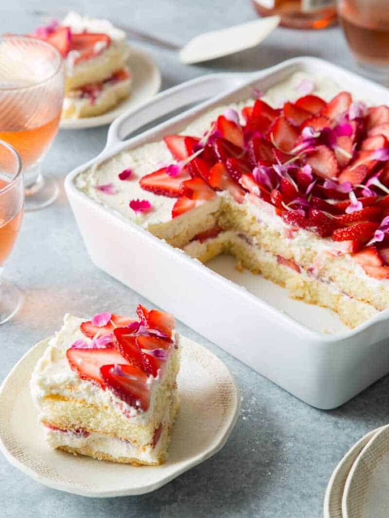 A baking pan of strawberry tiramisu with servings taken out on plates and drinks.