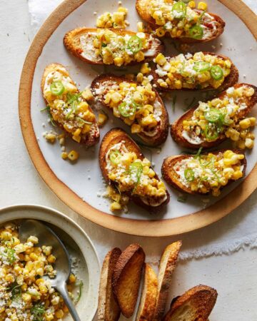 A plate of elote bruschetta with toppings and bread on the side.