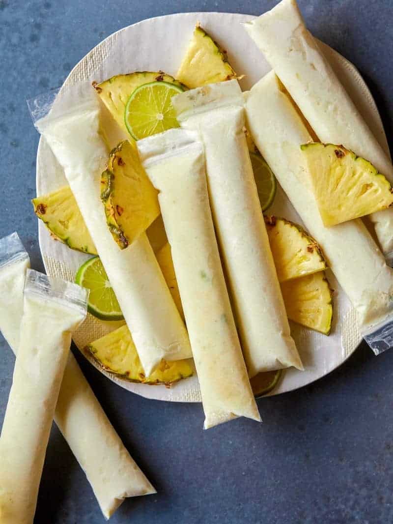 A plate of pina colada Italian ice pops with lime wheels and pineapple wedges.