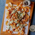 Homemade potato chips drizzled in gorgonzola cheese sauce on a wooden cutting board next to drinks.