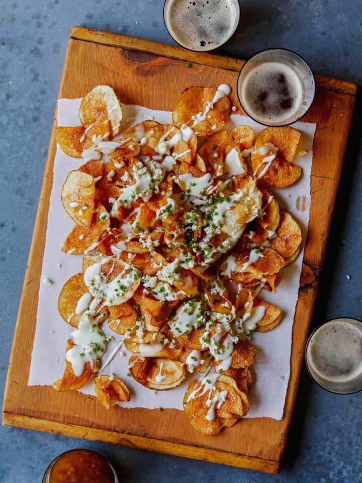 Homemade potato chips drizzled in gorgonzola cheese sauce on a wooden cutting board next to drinks.