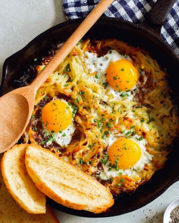 Simple cheesy skillet hash browns and eggs with bread and a wooden spoon.