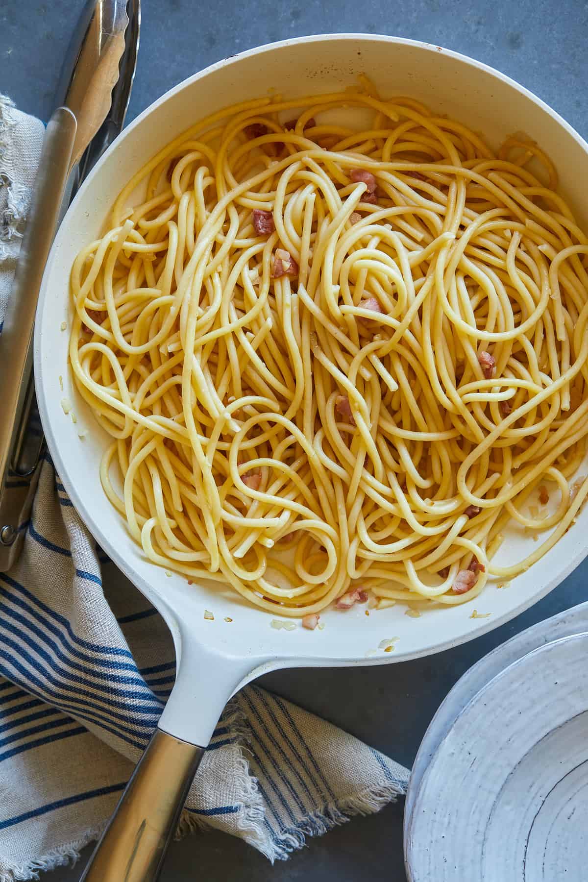 A pan of cooked bucatini and carbonara sauce and tongs.