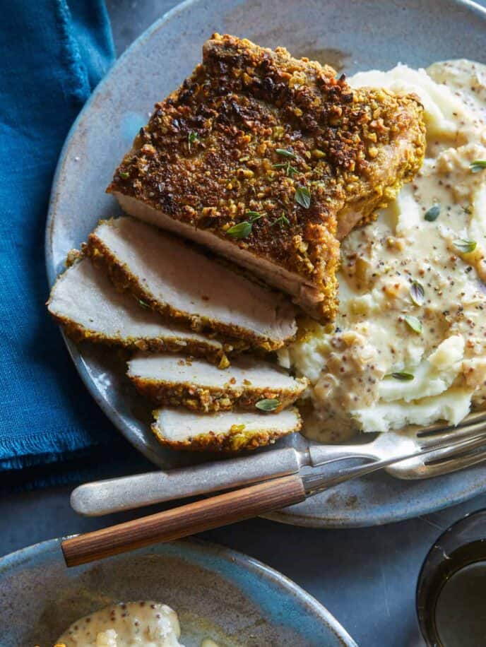 A close up of sliced pistachio crusted pork chop with mashed potatoes, a fork and knife.