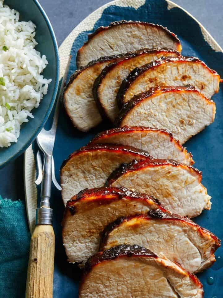 A platter of sliced char siu pork tenderloin next to white rice and a carving fork.