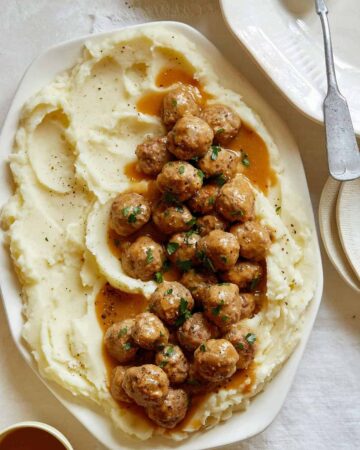 A platter of Swedish meatballs over mashed potatoes with plates to serve.