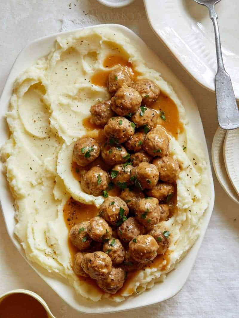 A platter of Swedish meatballs over mashed potatoes with plates to serve.