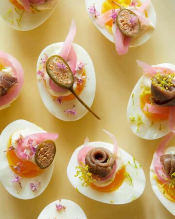 A close up of several undeviled eggs with pickled red onion, caperberries, and anchovies.