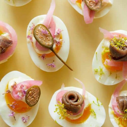 A close up of several undeviled eggs with pickled red onion, caperberries, and anchovies.