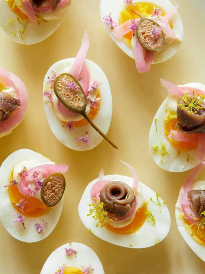 A close up of several undeviled eggs with pickled red onion, caperberries, and anchovies.