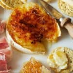 A close up of honey brûlée brie with crackers, meat, and cheese knife.