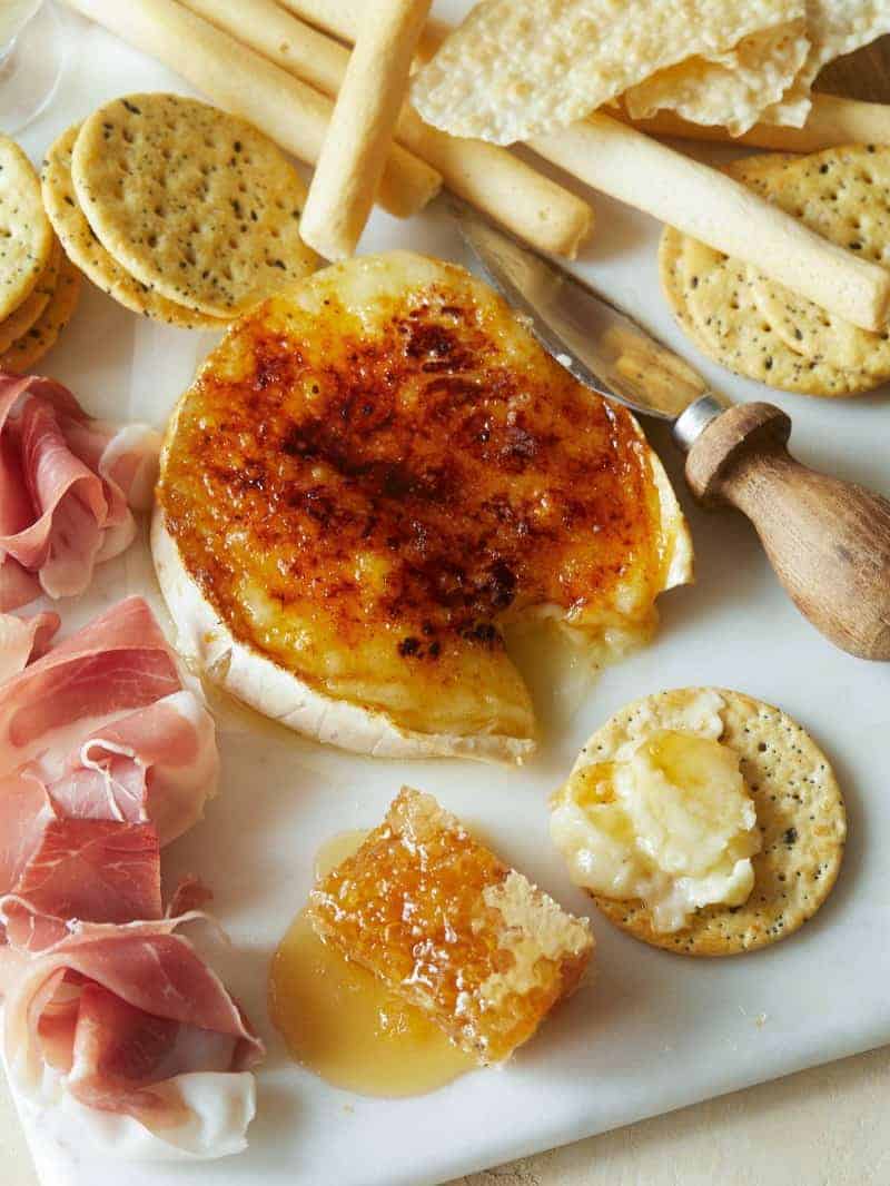 Honey Brûlée Brie on a platter with a piece taken out and spread on a cracker. The brie is surrounded by crackers and proscuitto. 