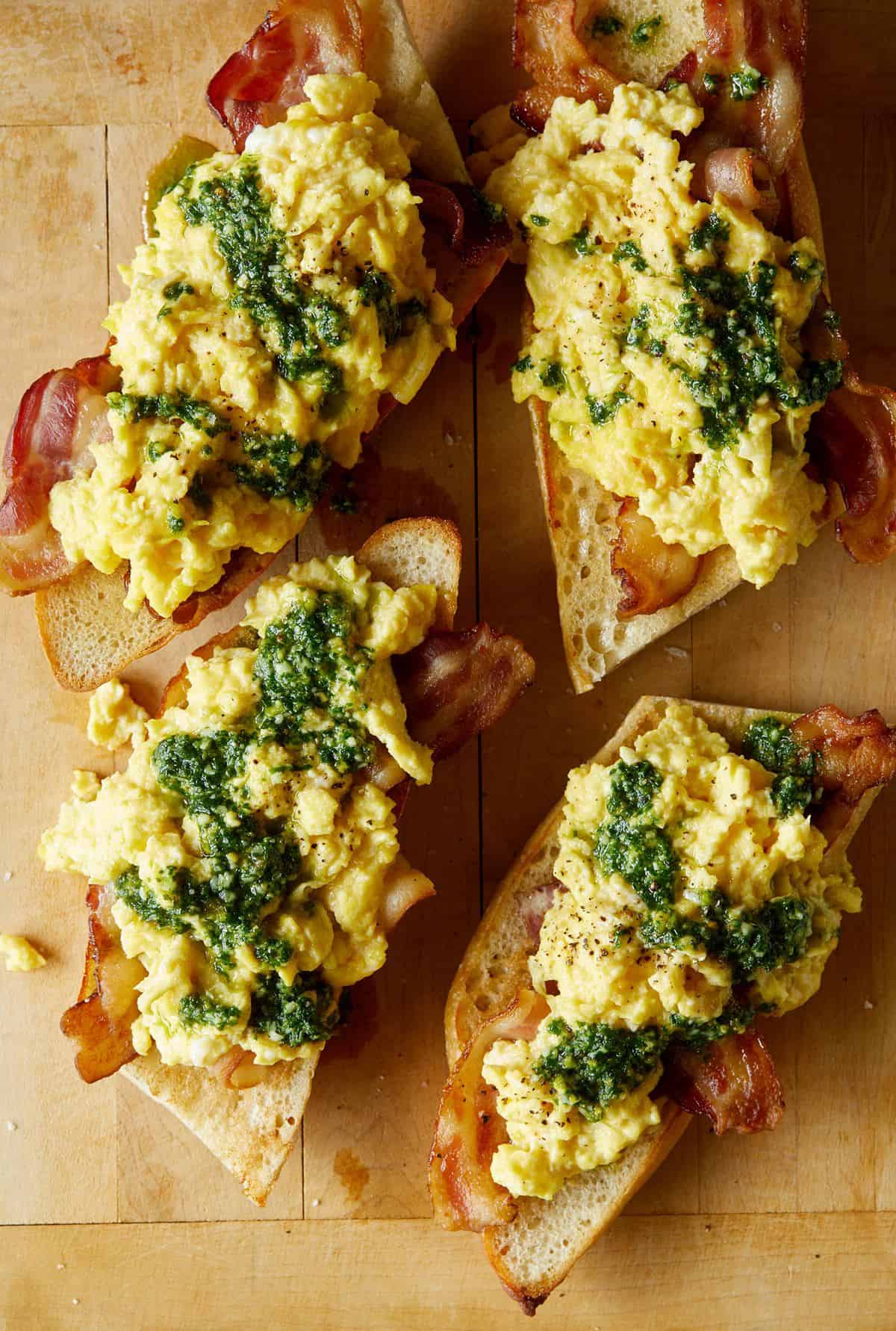 A close up of pesto topped scrambled eggs and bacon over toasted baguette.
