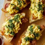 A close up of pesto topped scrambled eggs and bacon over toasted baguette.