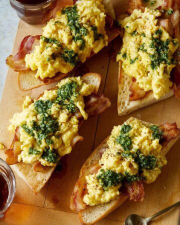 A close up of pesto topped scrambled eggs and bacon over toasted baguette.
