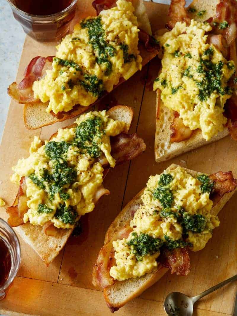 A close up of pesto scrambled eggs with bacon over toast on a wooden cutting board.