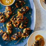 A platter of chicken satay with small plate servings and sauce on the side.