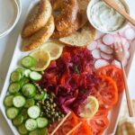 A platter of beet cured salmon, bagels and veggies with wooden serving utensils.