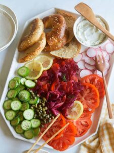 A platter of beet cured salmon, bagels and veggies with wooden serving utensils.