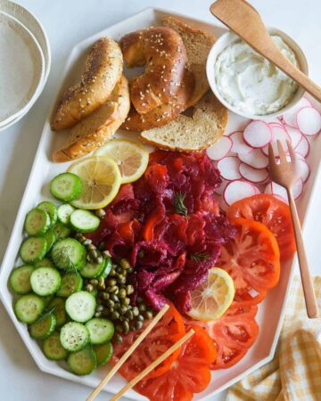 A platter of beet cured salmon, bagels and veggies with wooden serving utensils.