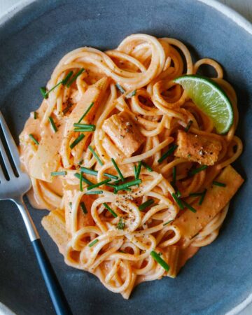 A close up of red curry noodles with a lime wedge and a fork.