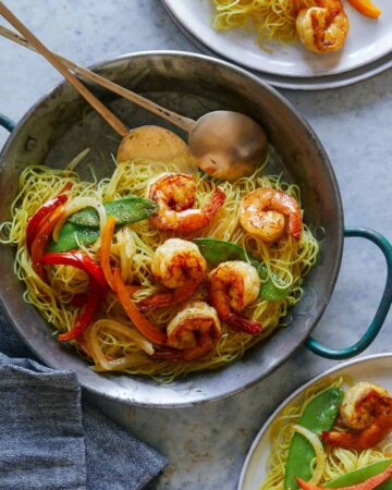 A pot of singapore noodles with shrimp and peppers and serving spoons.