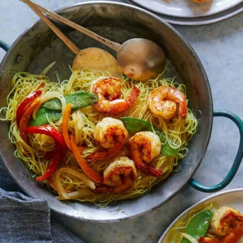 A pot of singapore noodles with shrimp and peppers and serving spoons.