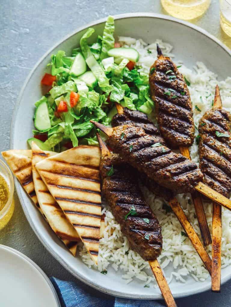 A plate of lamb kofta over basmati rice with veggies, grilled pita, and a drink.