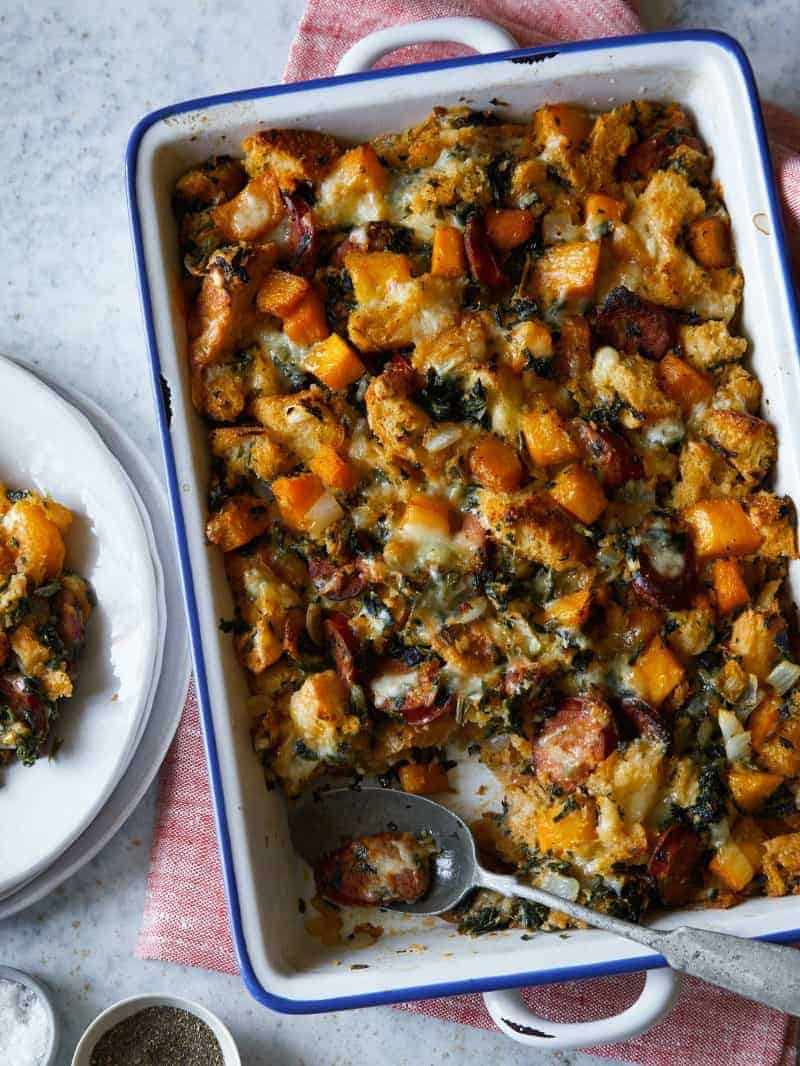 A baking dish of butternut squash, andouille, and herb stuffing with a spoon and plates.