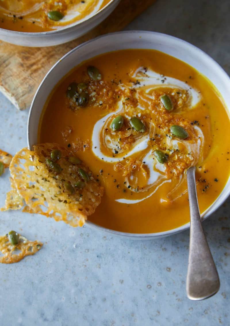 A bowl of creamy pumpkin soup with seeded parmesan crisps and a spoon.
