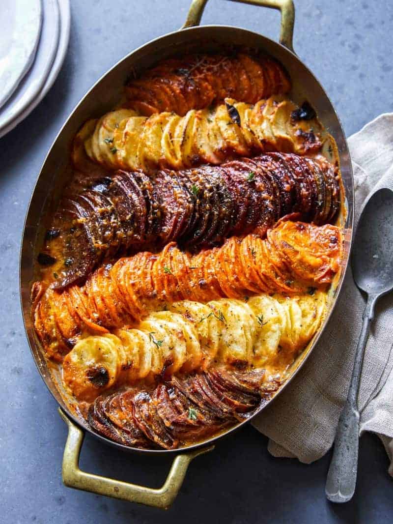 A pan of cheesy root vegetable gratin with linens and a spoon.