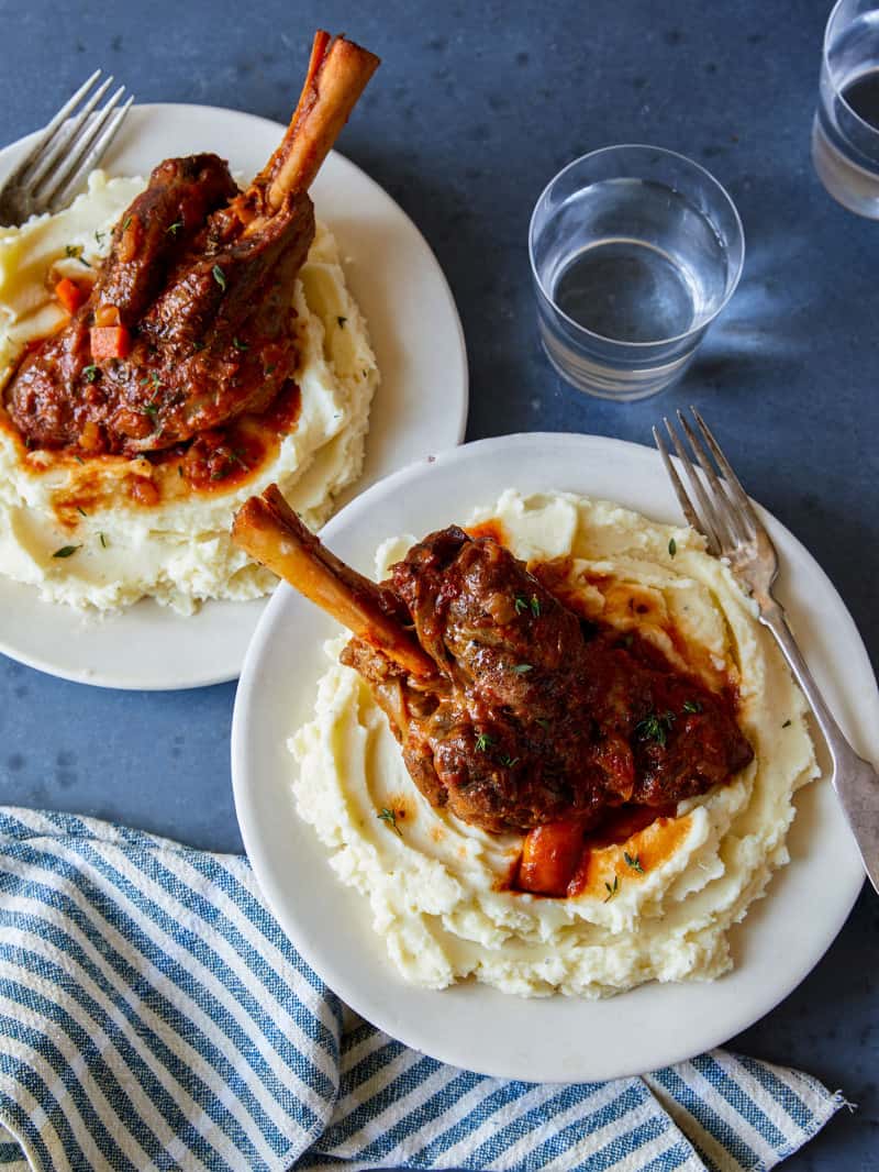 Plates of braised lamb shanks over creamy mashed potatoes with drinks, napkins, and forks.