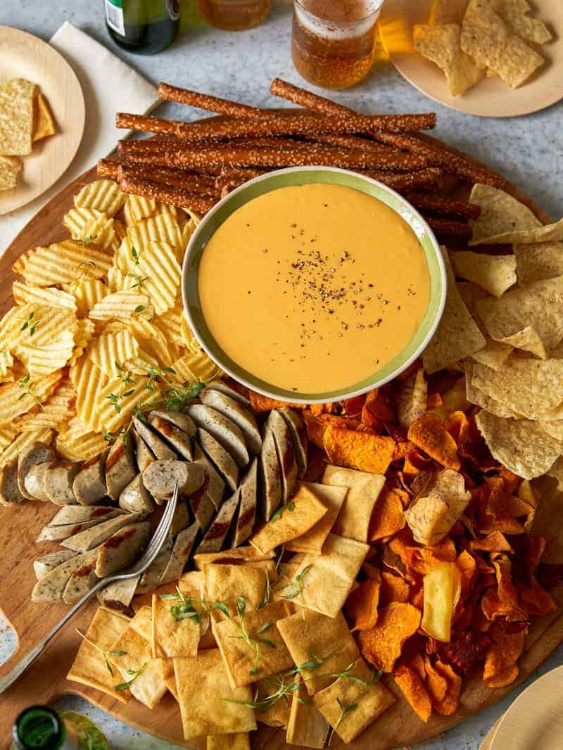 A bowl of crockpot beer cheese on a large platter of bratwursts, chips, and pretzel rods.