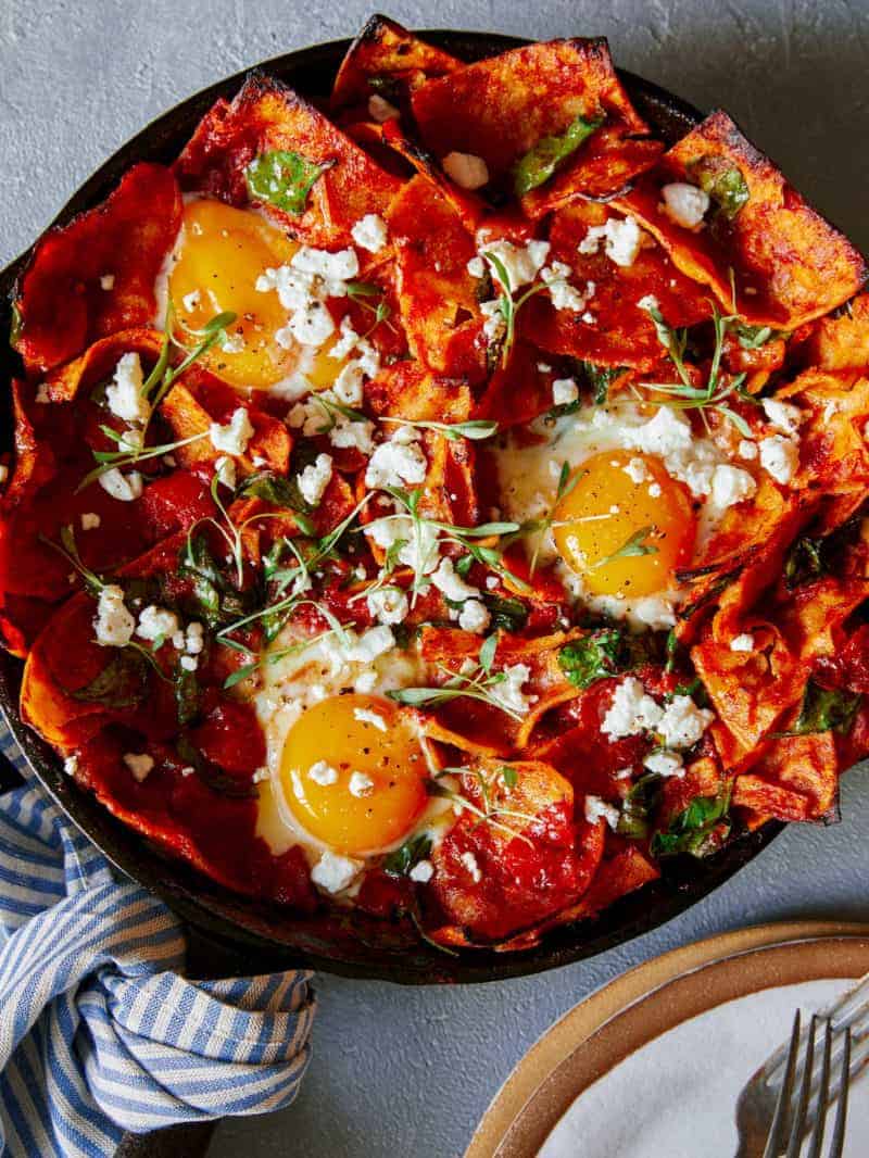 Shakshuka Chilaquiles in a skillet next to a stack of plates with forks. 