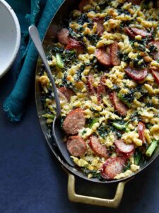 A close up of sausage kale and spaetzle pesto bake in a pan with a spoon.