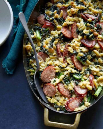 A close up of sausage kale and spaetzle pesto bake in a pan with a spoon.